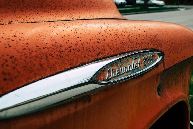 Close-up of a vintage Chevrolet 3600 emblem on a rusty orange truck outdoors.